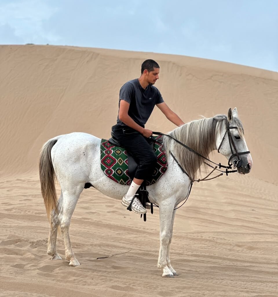 Male traveler riding a speckled white horse across desert sand dunes in Morocco