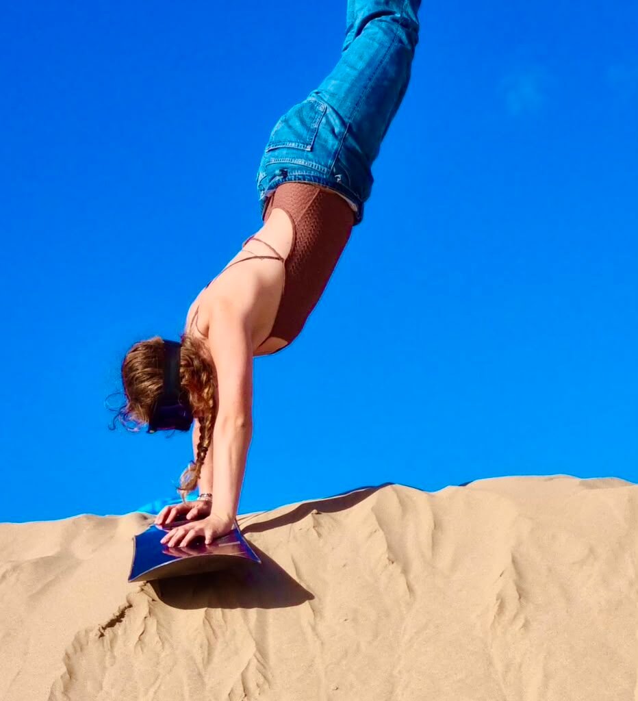 Athletic tourist doing a handstand on a sandboard atop a desert dune under a clear blue sky