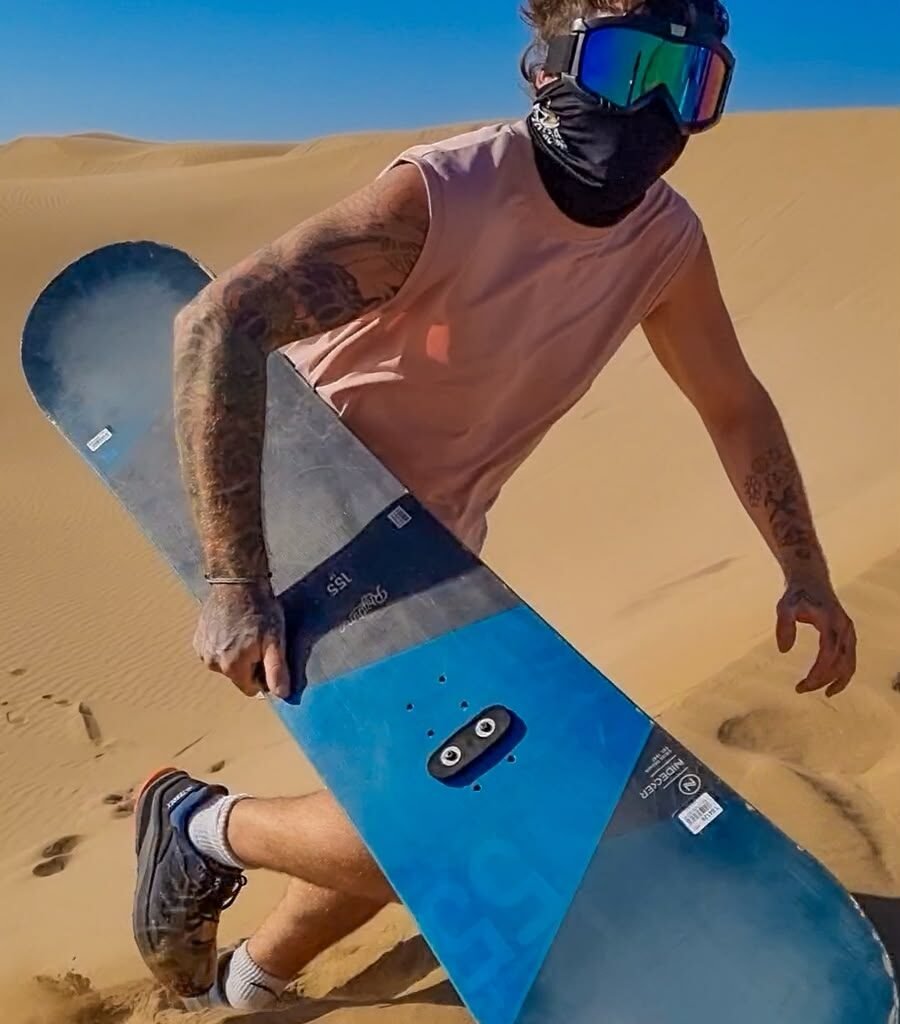 Man wearing protective goggles and face mask holding a sandboard on desert dunes