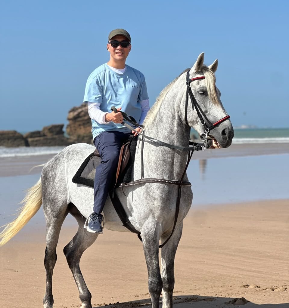 Male traveler in a cap and sunglasses riding a white horse along the beach