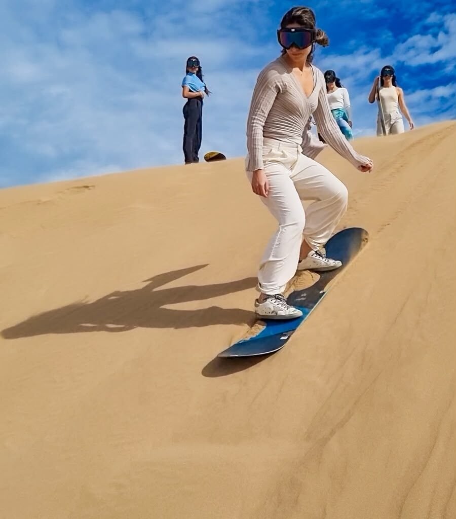 Woman sandboarding down a desert dune with friends watching from the top