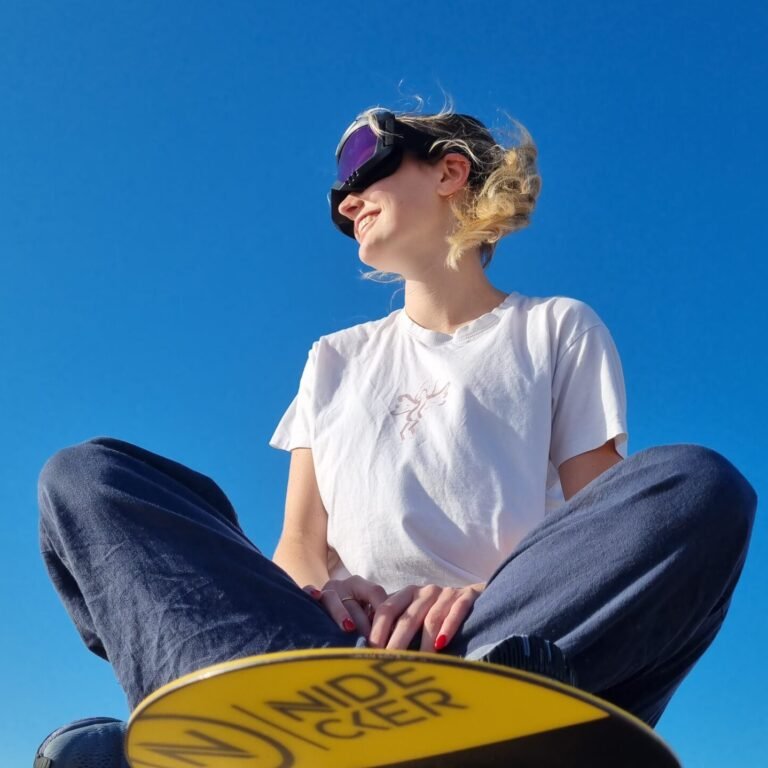 Woman wearing ski goggles sitting cross-legged on a sandboard atop a desert dune