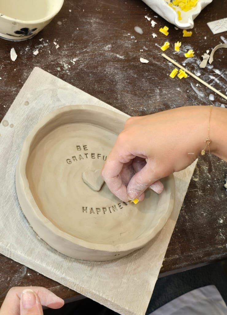 Person pressing yellow letter stamps into a handmade clay dish with the words "BE GRATEFUL HAPPINESS"