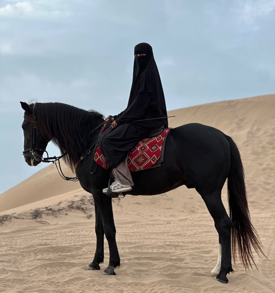 Woman wearing a black niqab riding a black horse across desert sand dunes in Morocco