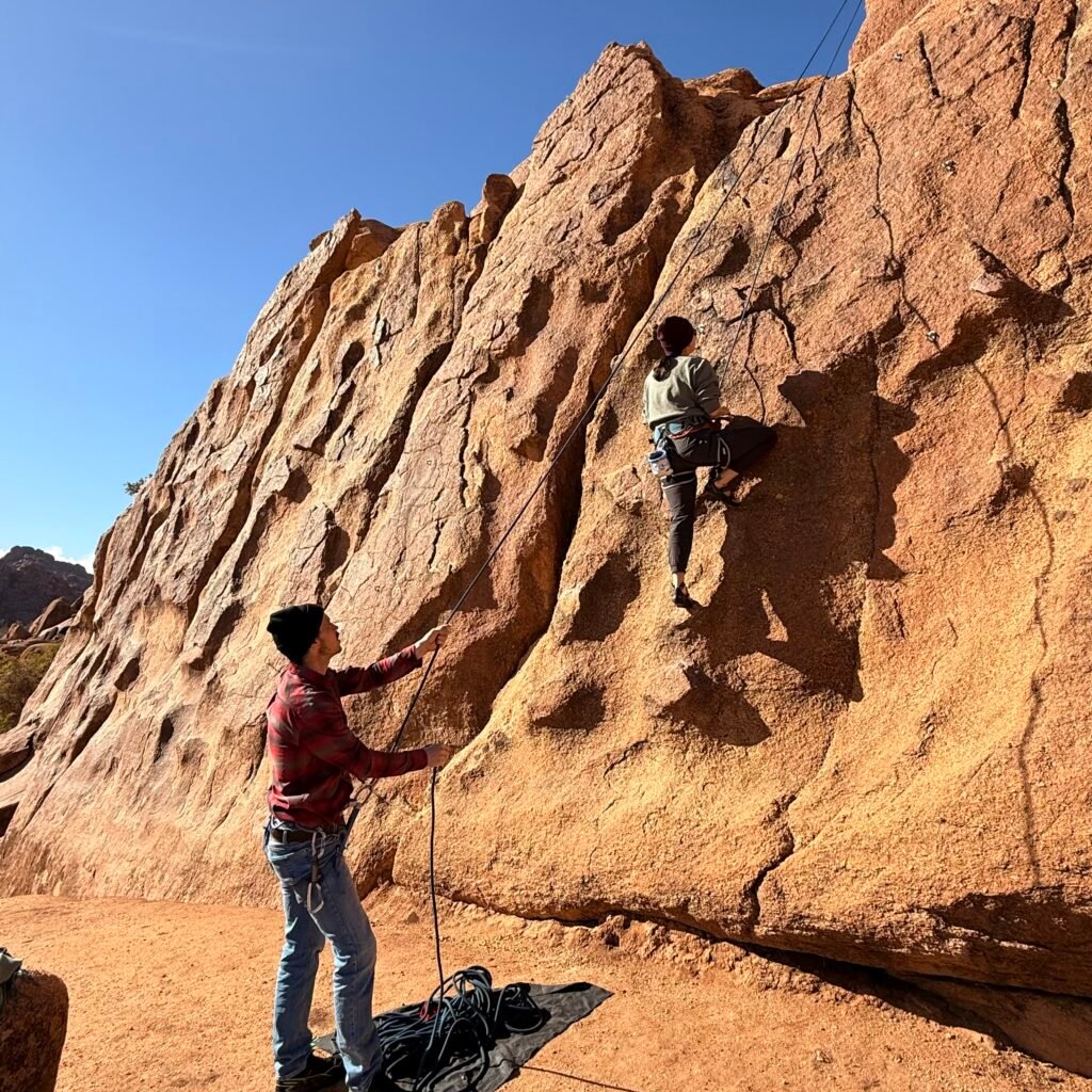 Female climber wearing a yellow helmet managing coiled rope at the base of a granite rock wall in Tafraoute, Morocco