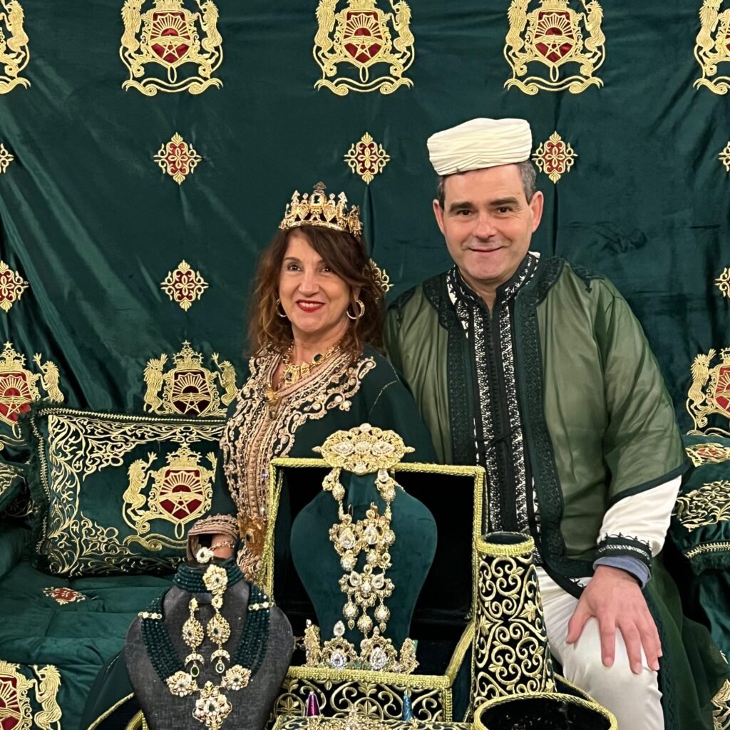 A couple posing in traditional Moroccan wedding attire in front of a green decorative backdrop