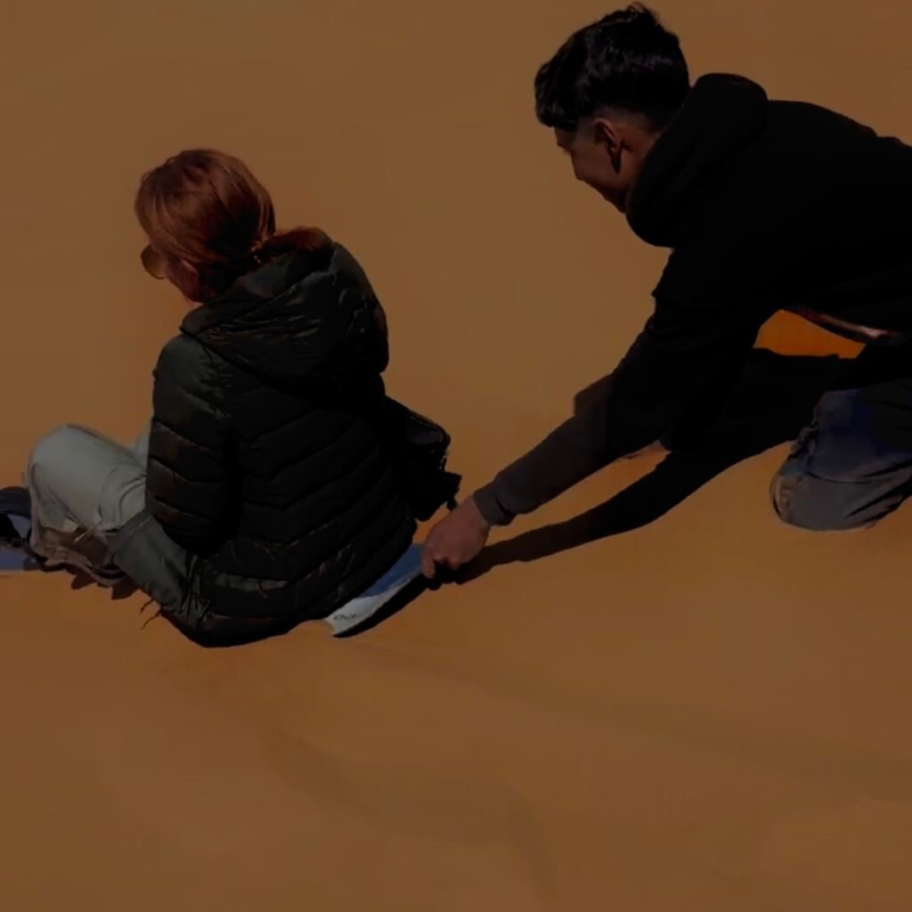 Tourist sandboarding down a desert dune assisted by a guide