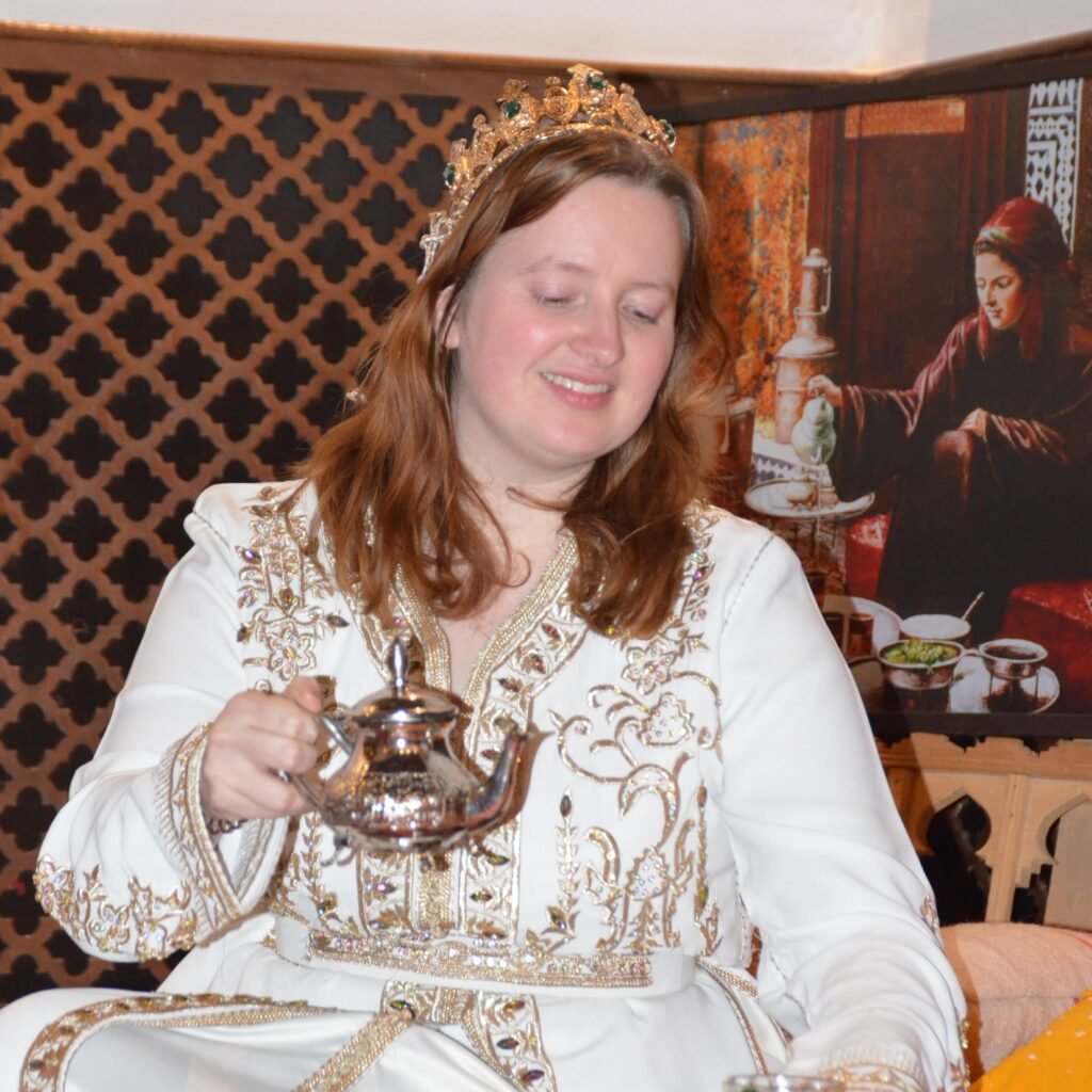 A female traveler wearing a gold tiara and embroidered white caftan pouring mint tea during a traditional Moroccan ceremony