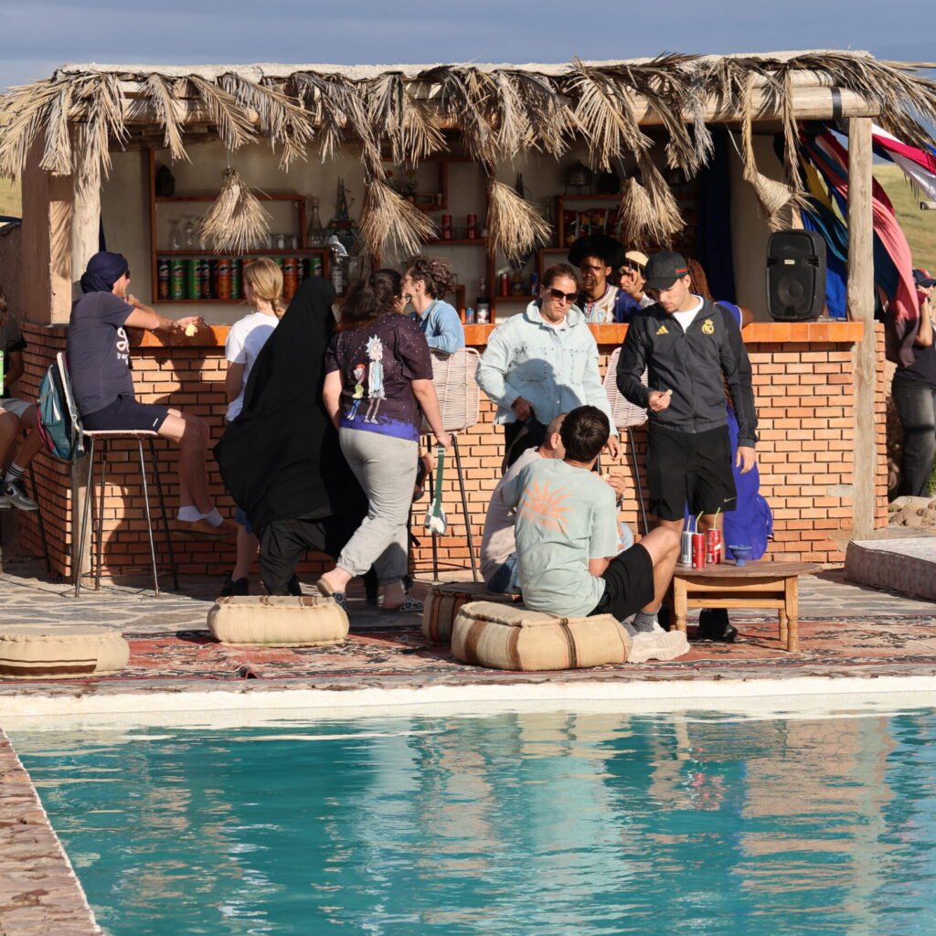 Travelers relaxing by a pool near a thatched-roof outdoor bar during a Moroccan tour