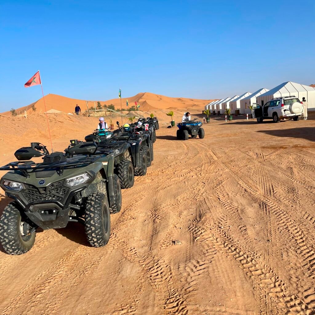 A line of quad bikes parked along a sandy track outside luxury desert camp tents under a clear blue sky