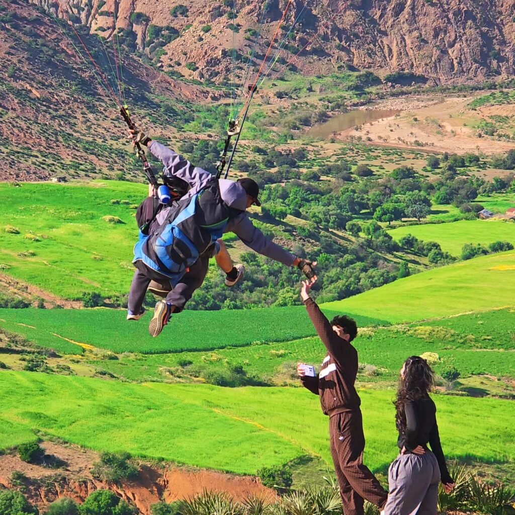 Paraglider high-fiving a spectator over a green valley and mountains