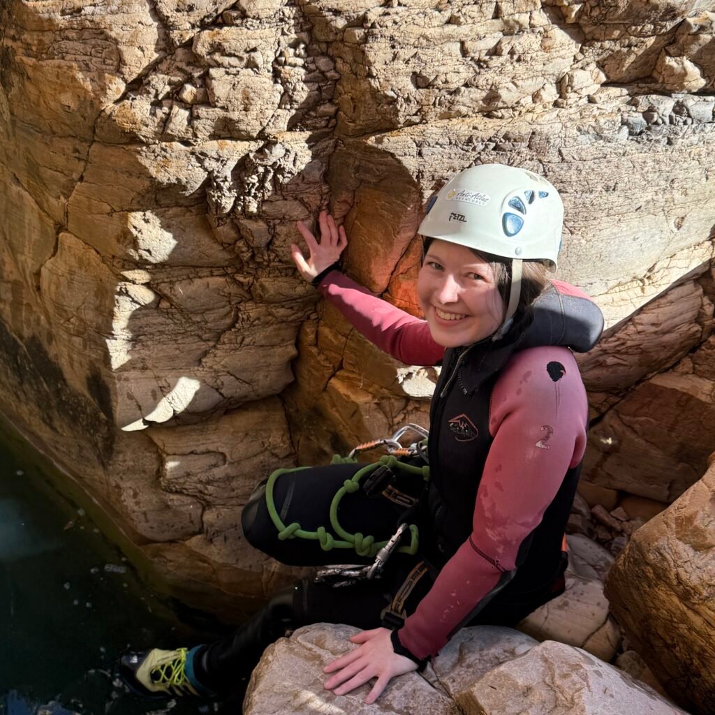 Female traveler in a helmet and wetsuit leaning against a rocky canyon wall above a natural pool in Morocco