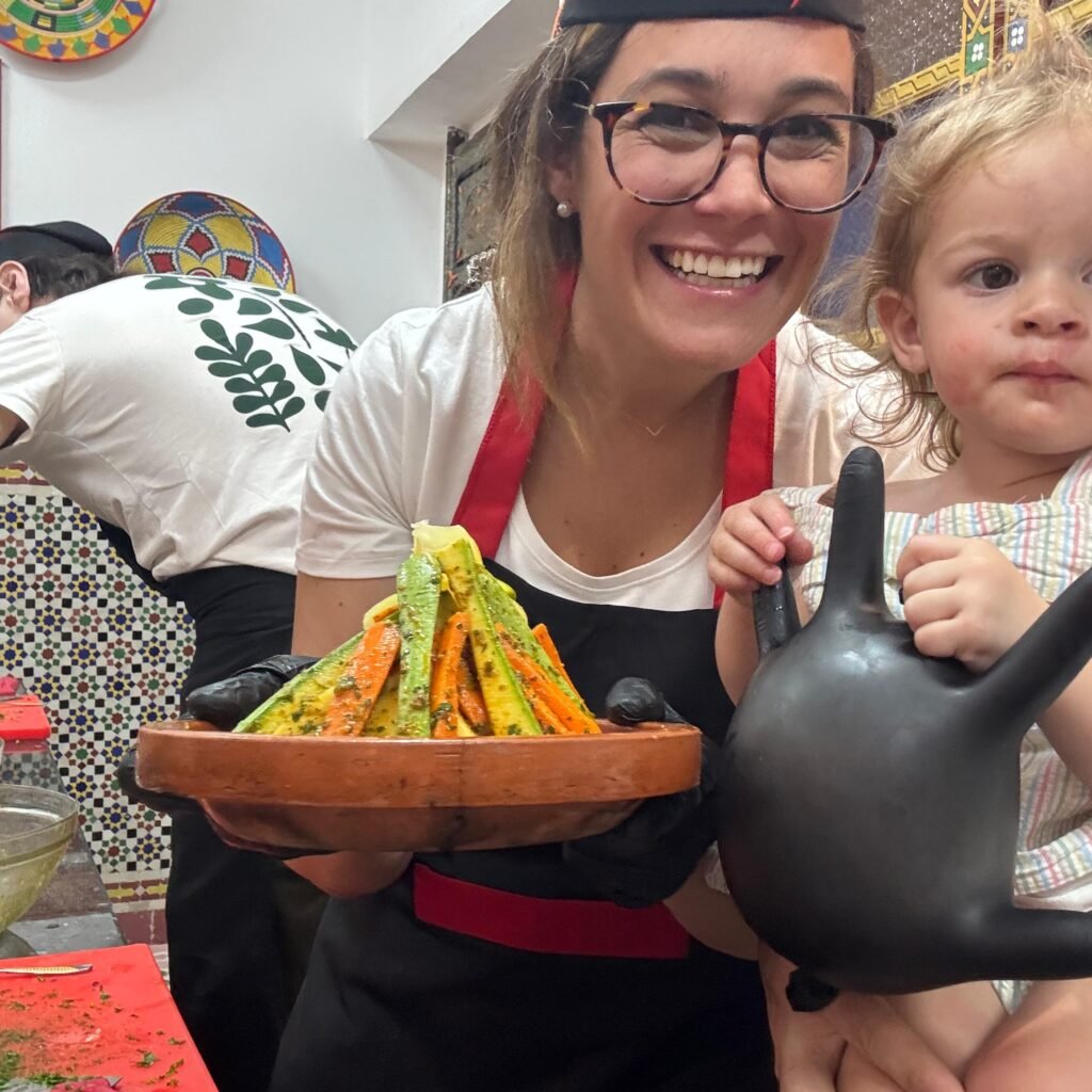 Smiling female chef holding a prepared tagine dish with colorful vegetables alongside a child in a kitchen