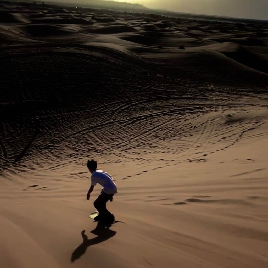 Person sandboarding down a steep desert sand dune at sunset
