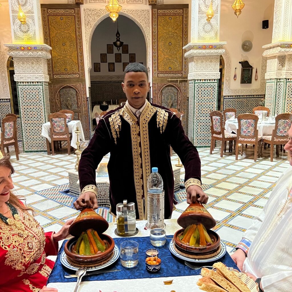 A waiter serving traditional vegetable tajines to a couple dressed in Moroccan wedding attire inside an ornate riad