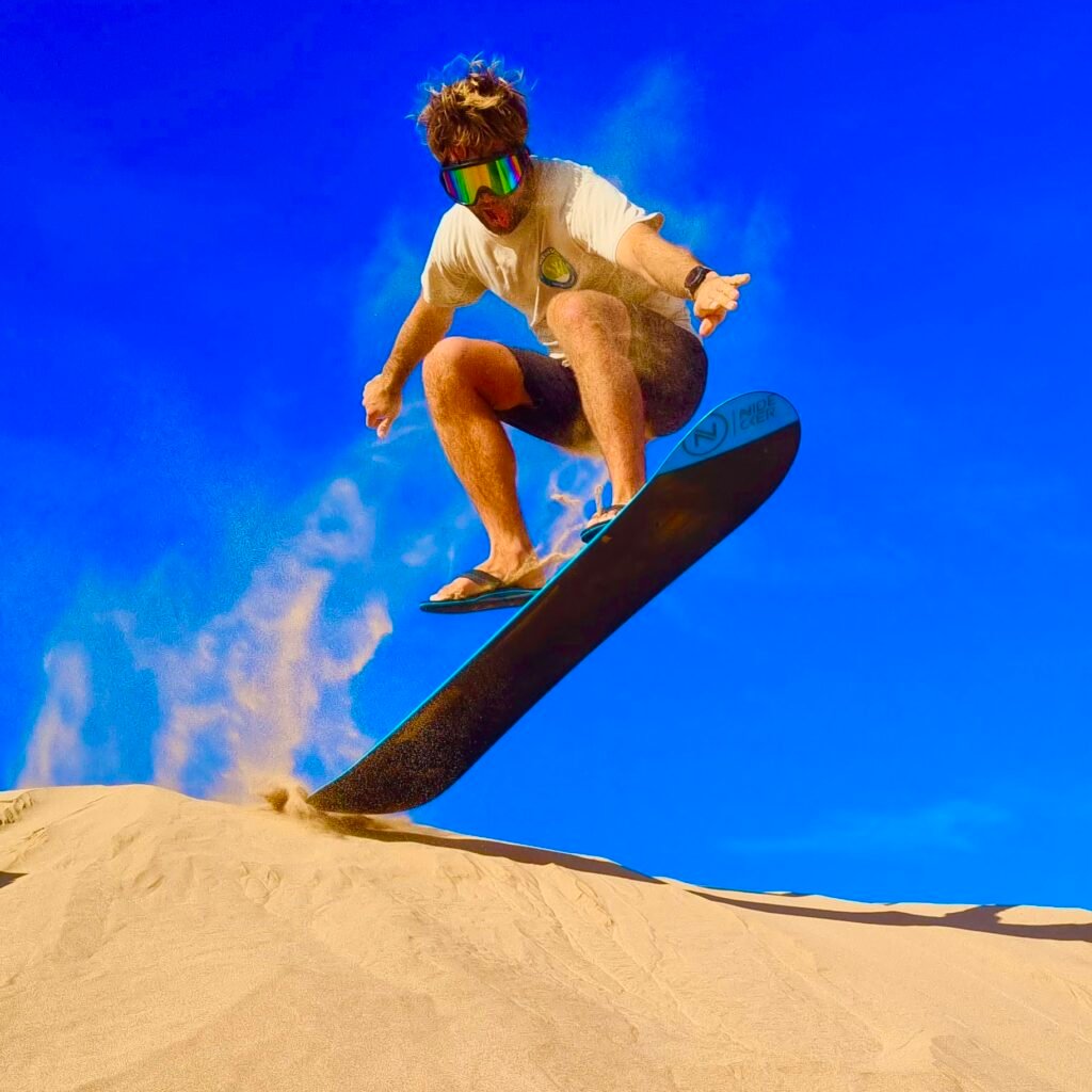 An adventurous traveler doing a high jump on a sandboard off a sand dune ridge under a vibrant blue sky