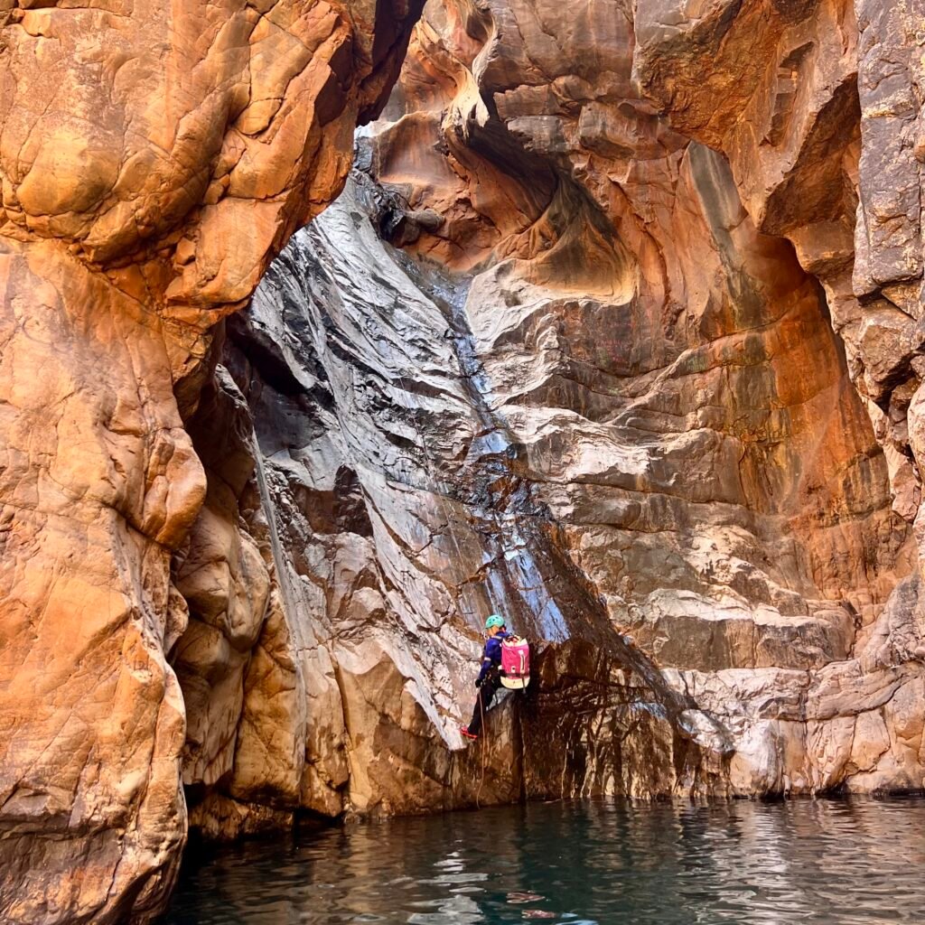 Traveler rappelling down a steep, dark rock face beside a waterfall into a deep canyon pool in Morocco