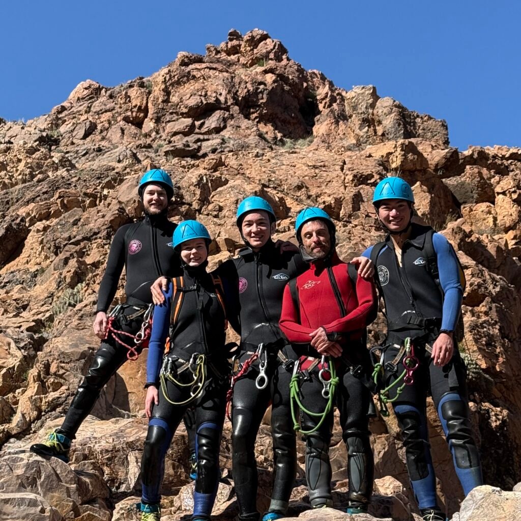 Group of five travelers in wetsuits and helmets posing against a rocky mountain backdrop in Morocco