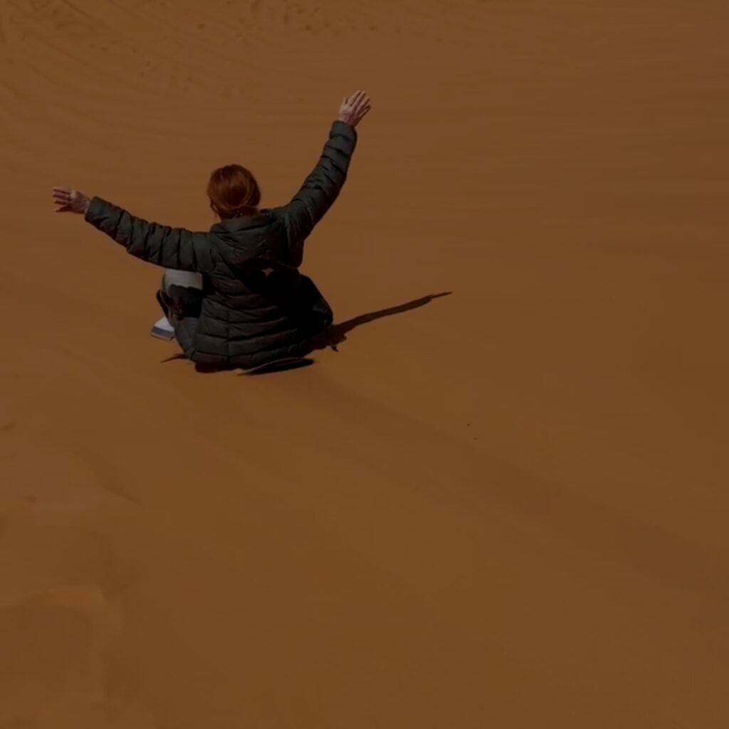 Person sliding down a high desert sand dune on a sandboard with open arms