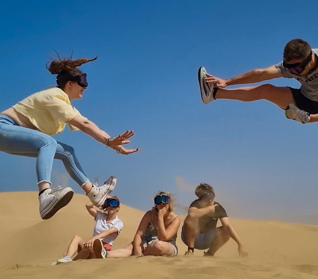 Group of energetic travelers jumping in mid-air during a Sahara desert sandboarding excursion