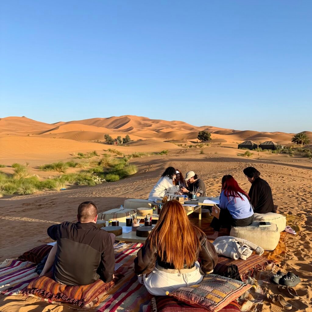 Group of people seated outdoors on traditional rugs and cushions in the Sahara Desert, participating in a workshop surrounded by sand dunes