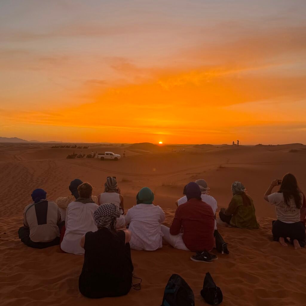 Group of travelers sitting on Sahara sand dunes watching a vibrant golden sunset