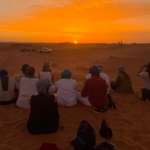 Group of travelers sitting on Sahara sand dunes watching a vibrant golden sunset
