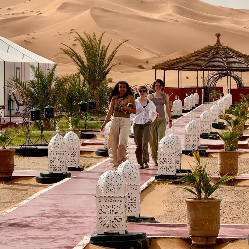 Group of travelers walking through a Sahara desert camp with towering sand dunes in the background