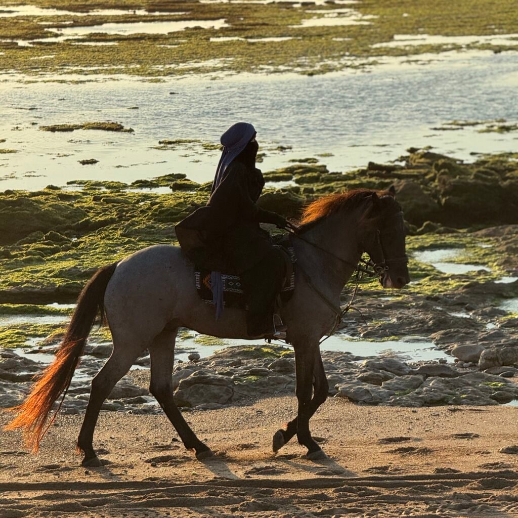 Traveler in a black robe riding a horse along a sandy beach near tide pools at sunset in Morocco