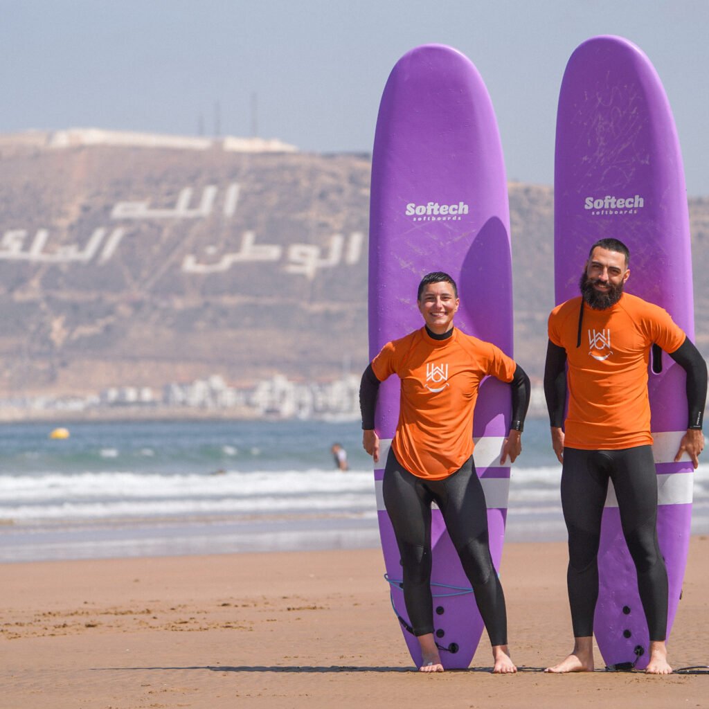 Two surfers standing with purple softboard surfboards on Agadir beach with the hillside in the background