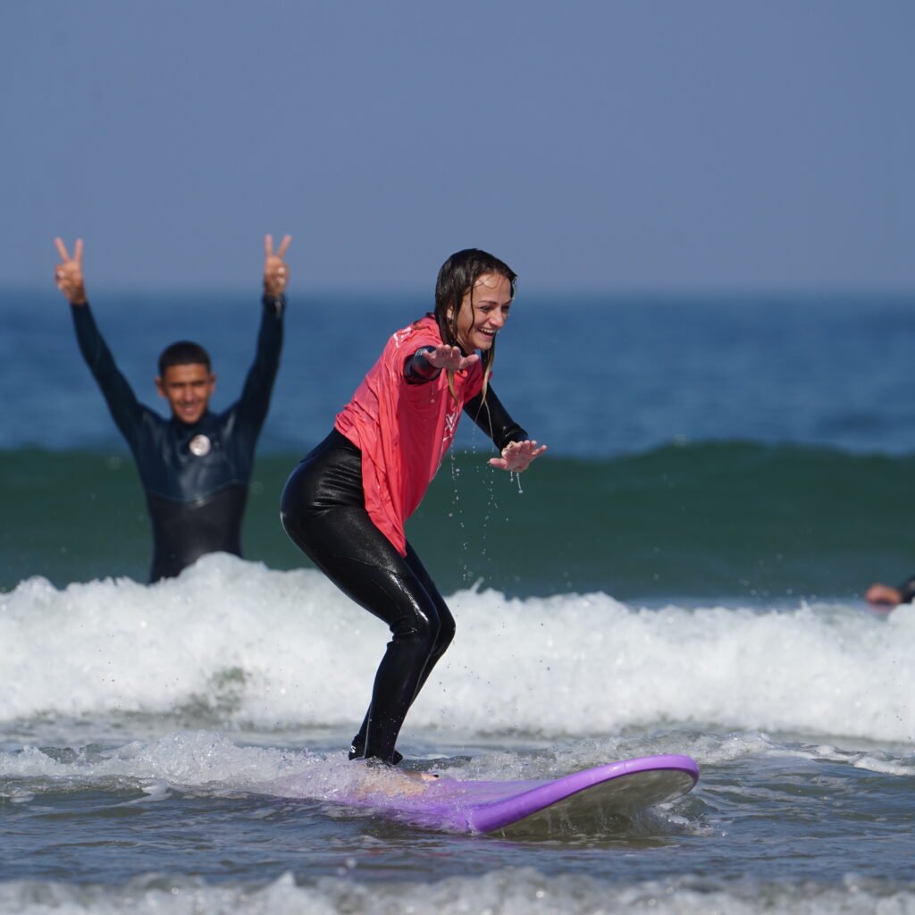 Female surfer riding a small wave on a purple surfboard with a fellow surfer in the background