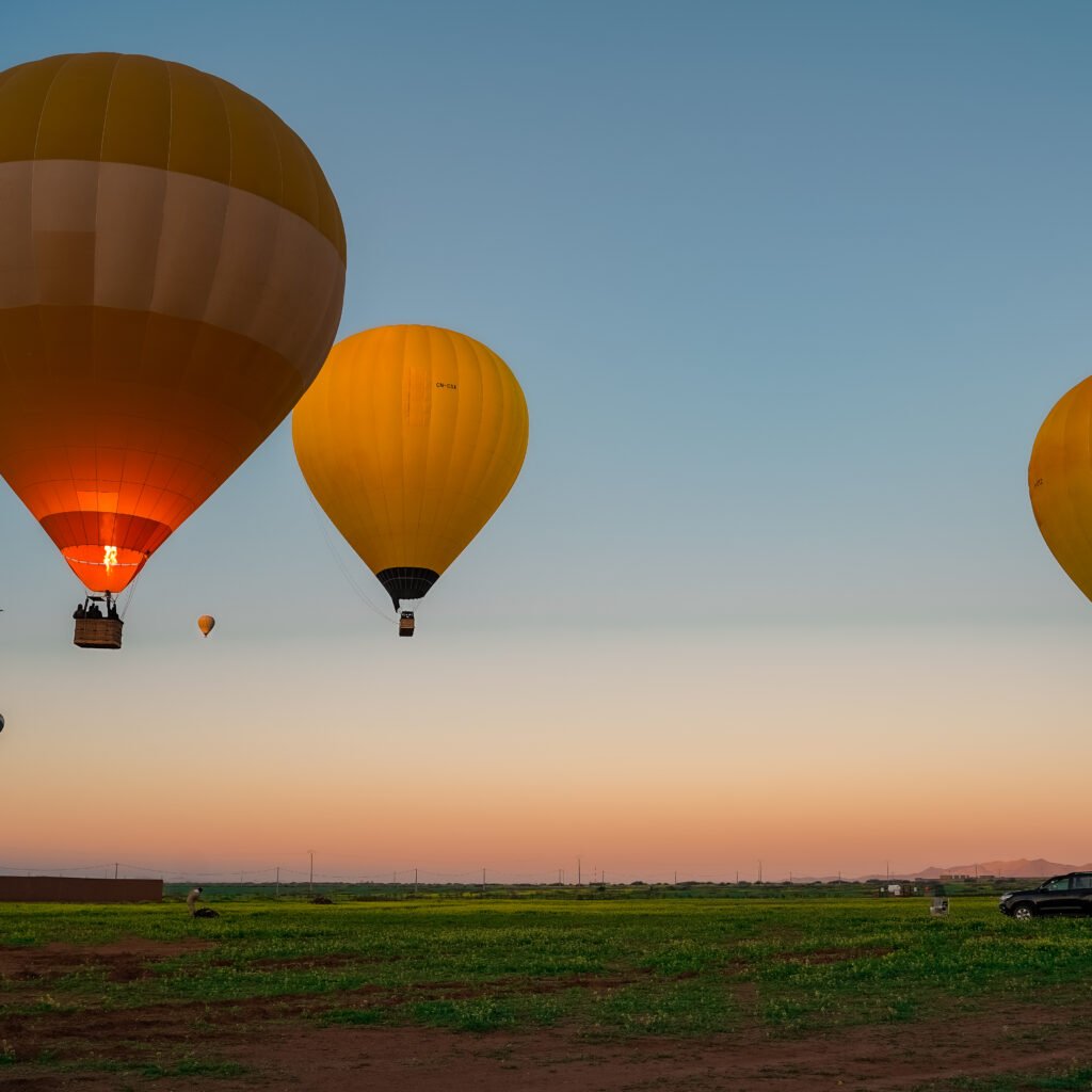 Colorful hot air balloons glowing with burner flames floating over a green field at sunrise with mountains in the background.