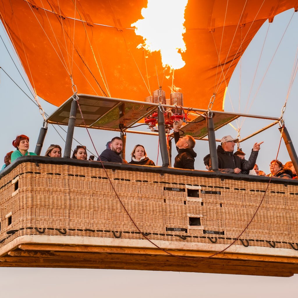 Passengers enjoying a hot air balloon ride in a large wicker basket as the burner fires a bright flame into the orange envelope against a clear sky.