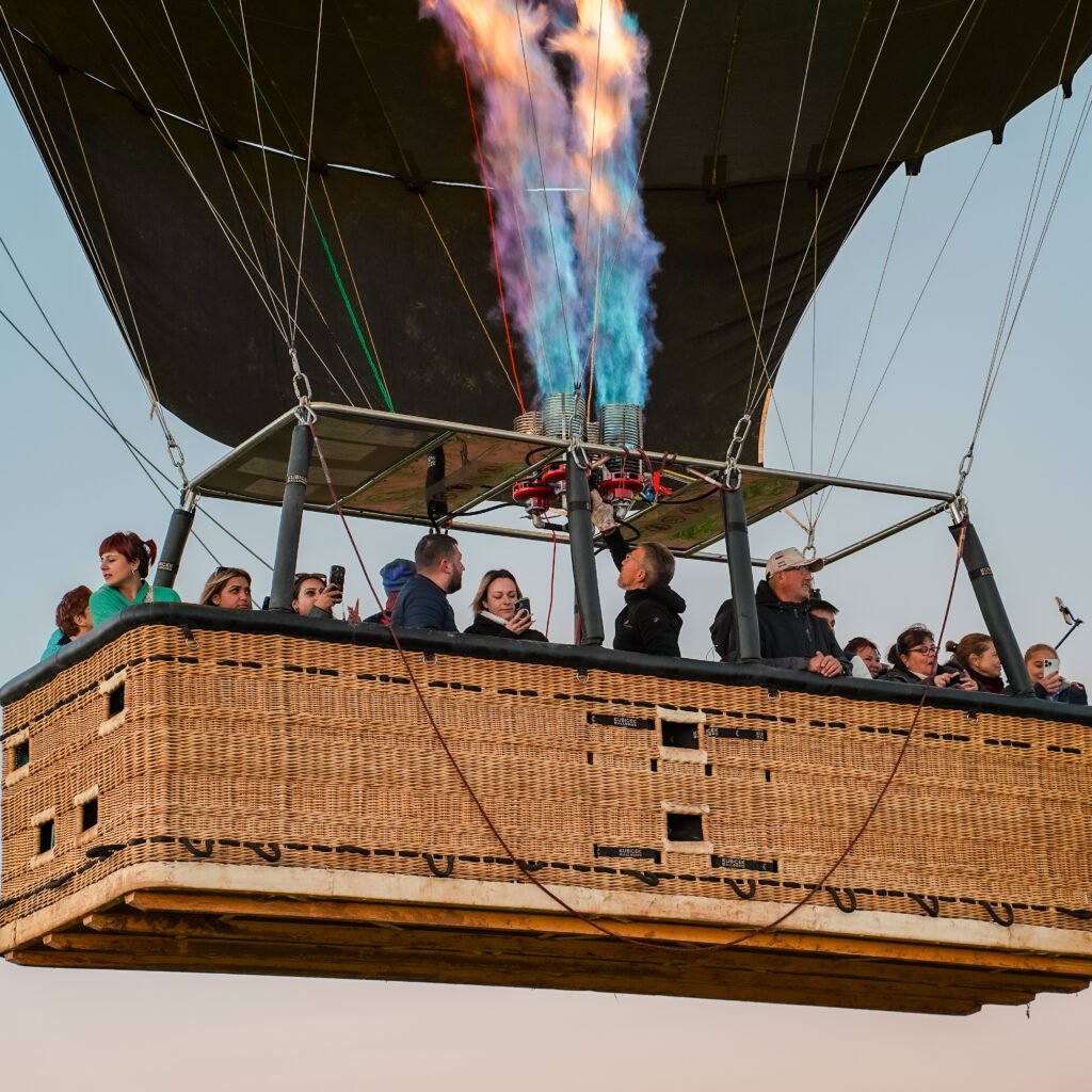 Passengers in a large wicker hot air balloon basket watching the burner fire a spectacular blue and orange flame into the envelope during a scenic flight.
