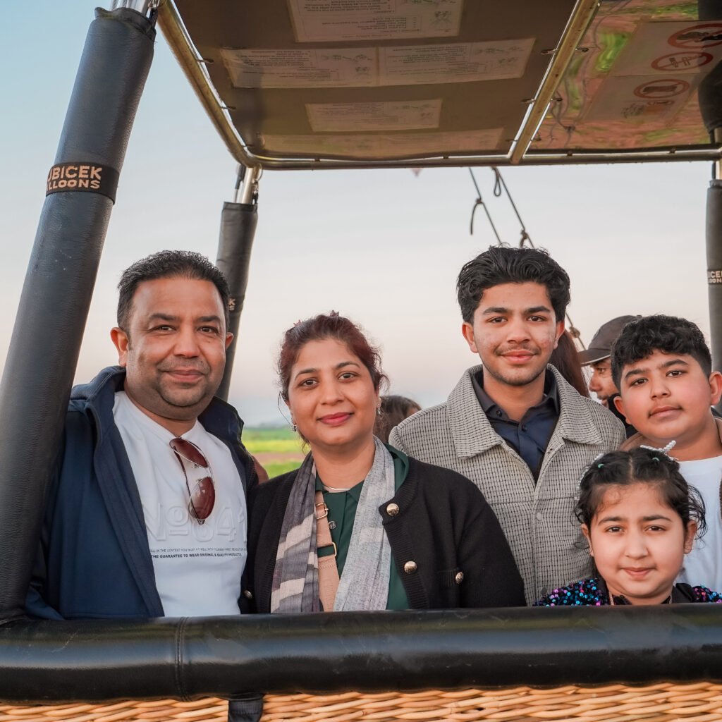 Happy family of five posing together in a Kubicek hot air balloon basket with green fields and another balloon visible in the background at sunrise.