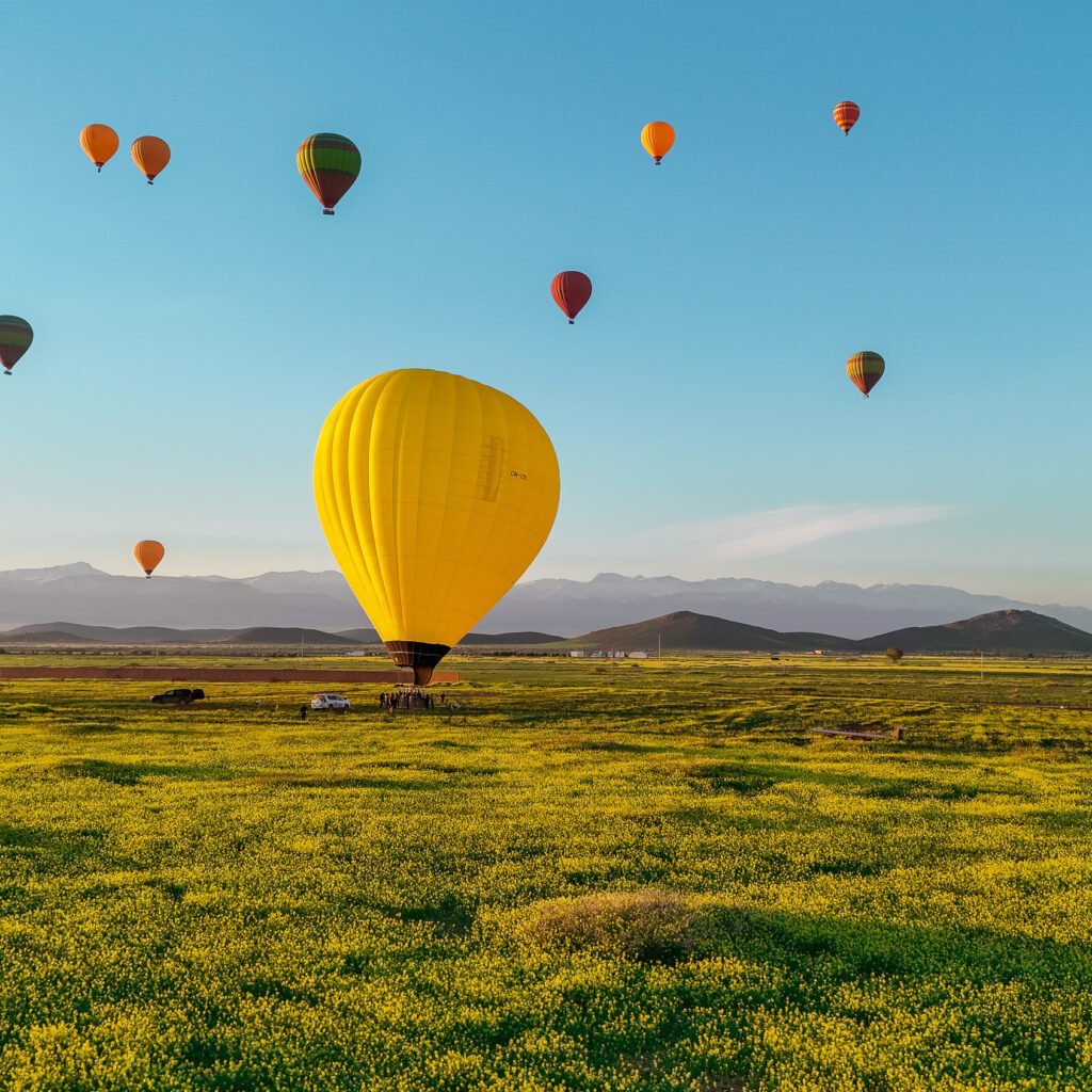 Yellow hot air balloon landing in a green field with colorful balloons and mountains in background