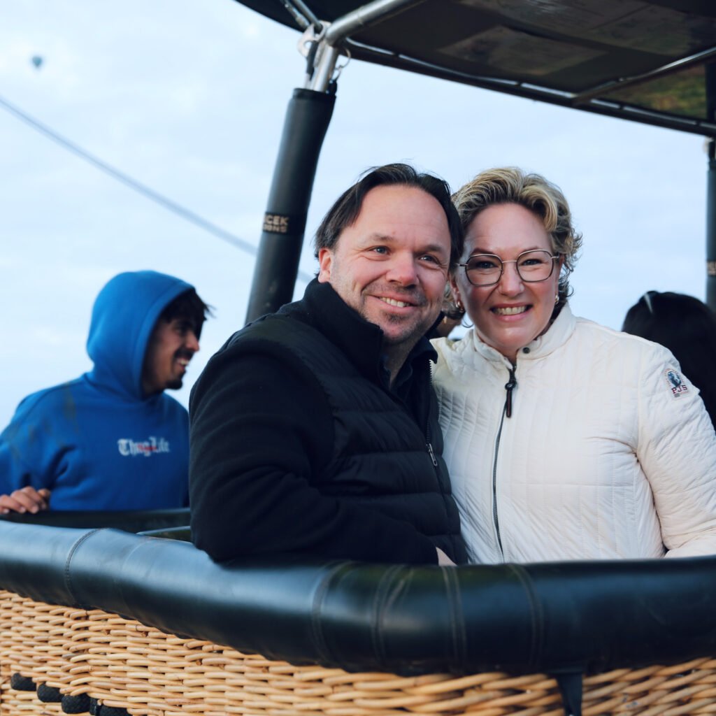 Smiling couple enjoying a hot air balloon ride in a wicker basket over a scenic landscape.