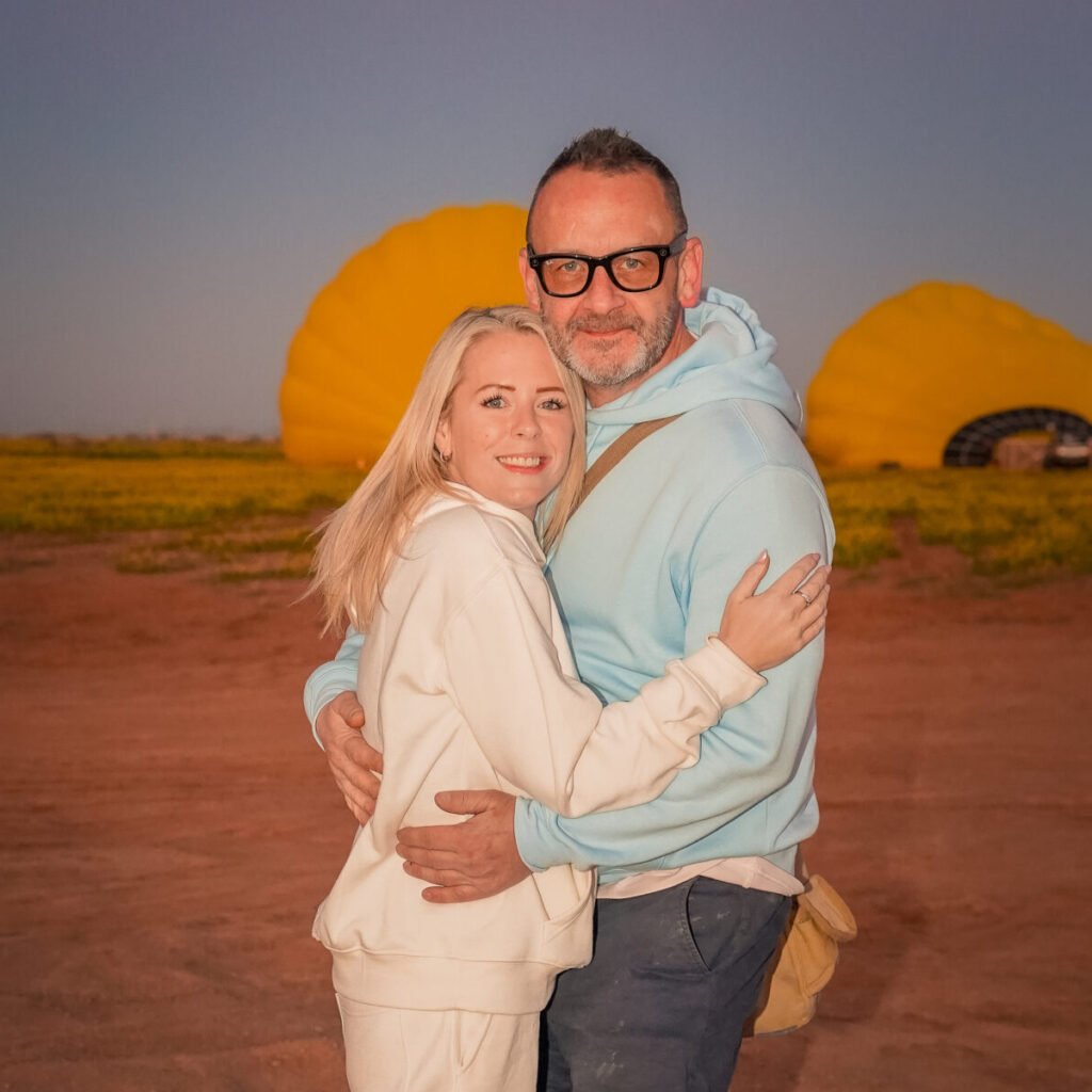 Happy couple embracing in front of colorful hot air balloons being inflated on the ground at sunrise.