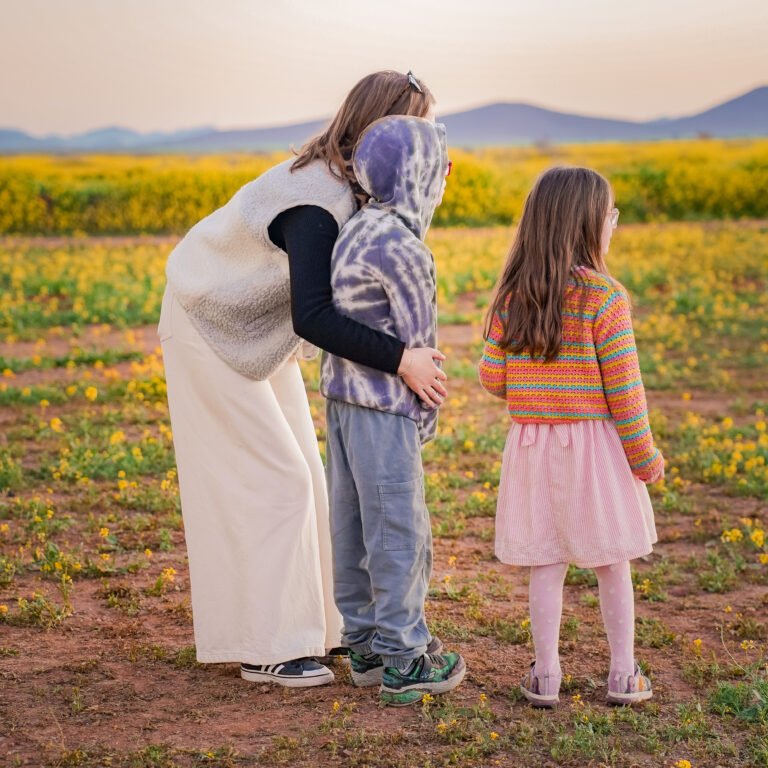 Mother and two children standing in a field of yellow flowers watching hot air balloons at sunrise with mountains in the background.