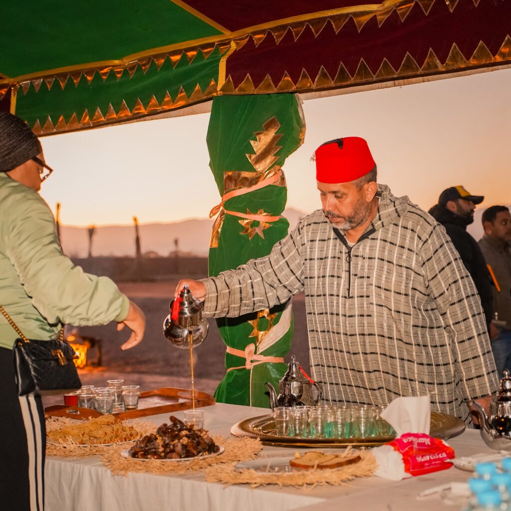 Man in traditional Moroccan attire pouring mint tea for guests at an outdoor tent reception with a scenic sunrise and mountains in the background.