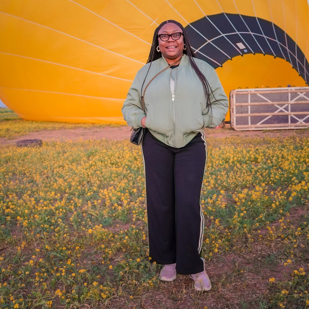 Happy woman posing in a field of yellow flowers in front of a large yellow hot air balloon being inflated at sunrise.
