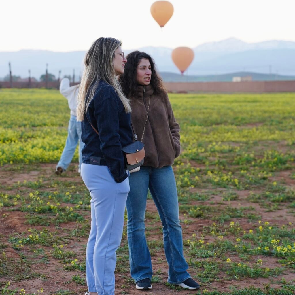 Two women standing in a field of yellow flowers watching colorful hot air balloons float over the landscape with mountains in the background at sunrise.