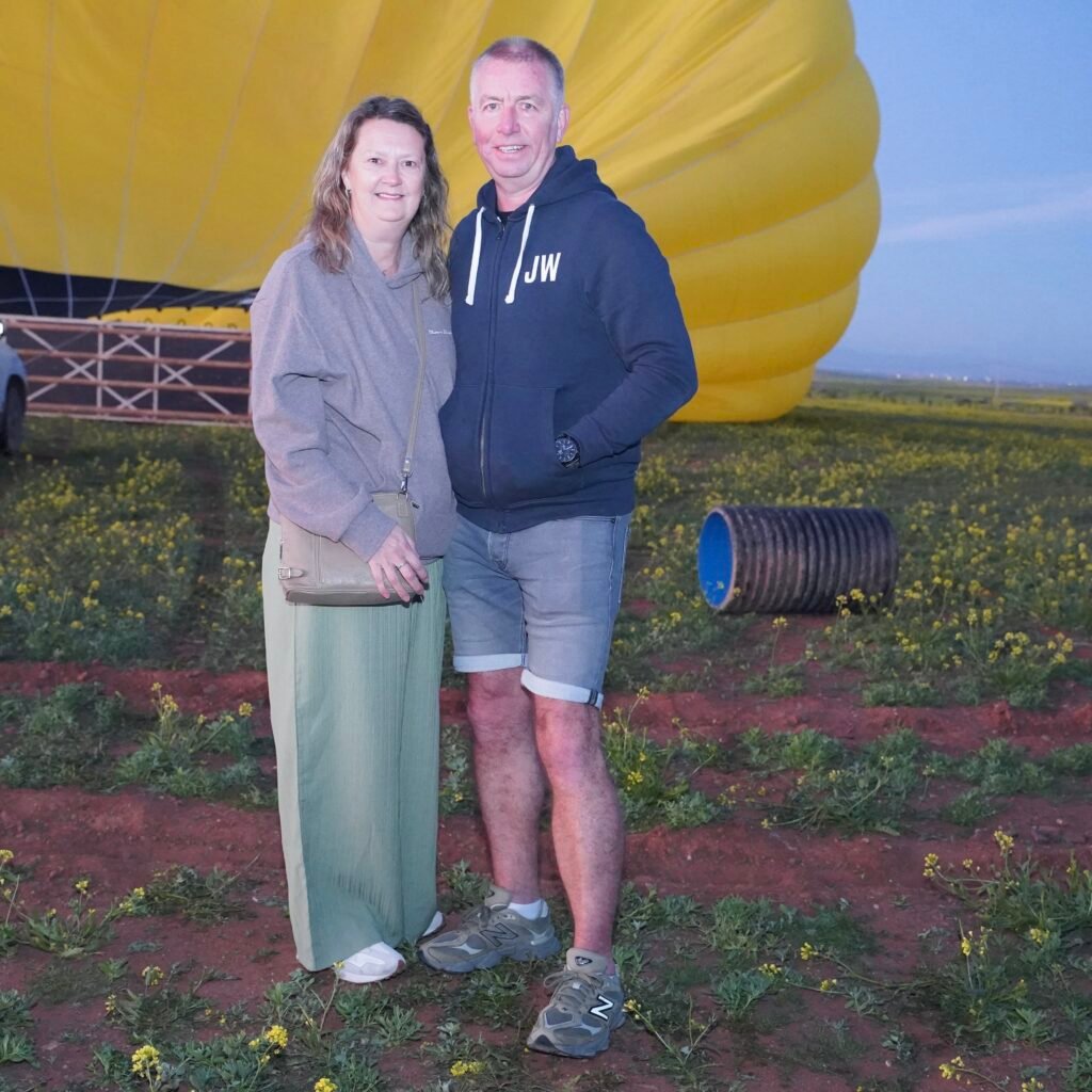 Couple posing together in a field of yellow flowers with a large yellow hot air balloon and support vehicle in the background at sunrise.