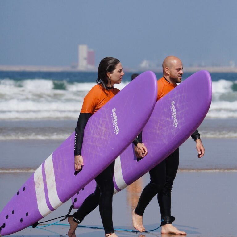 Man and woman walking along the wet sand carrying purple softboard surfboards