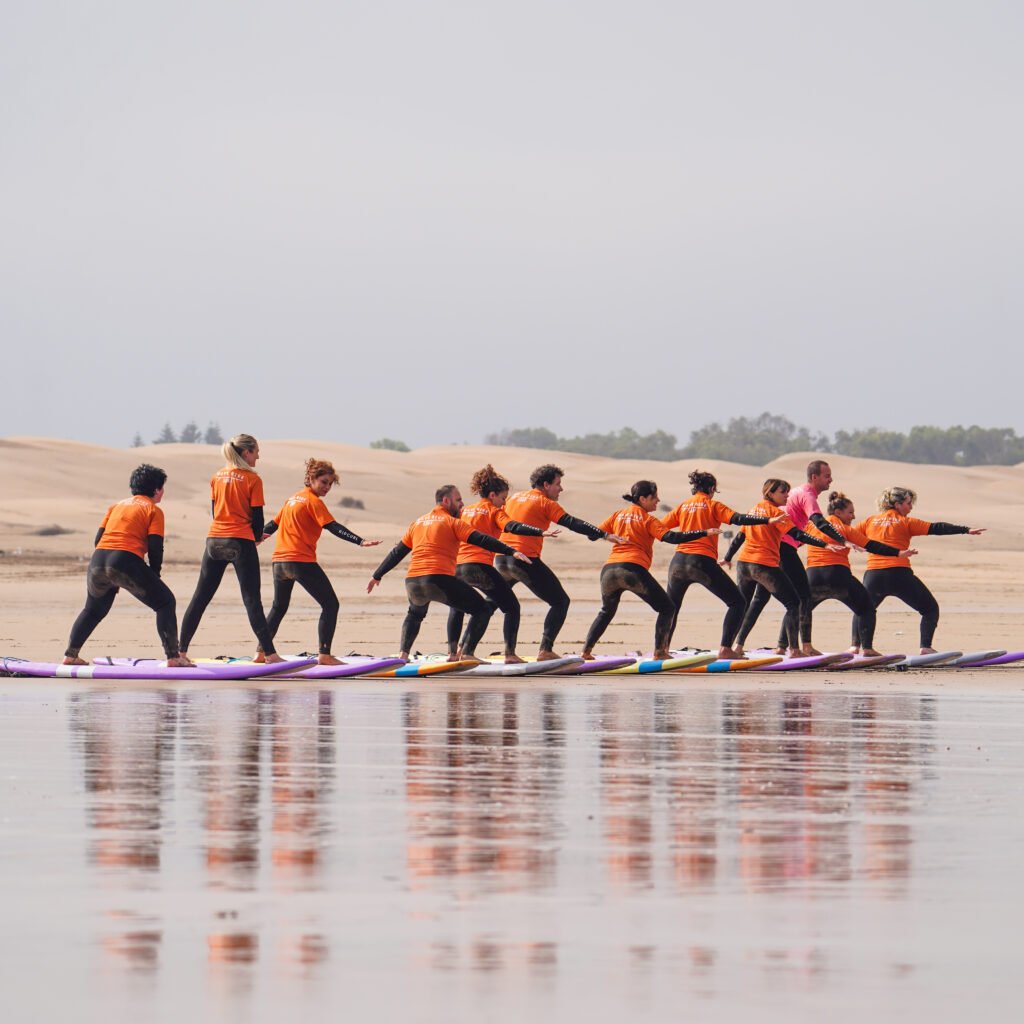 Group of surf students in matching orange tops practicing stance on surfboards line on the beach