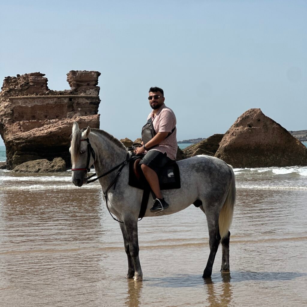 Male traveler riding a white horse in the ocean tide near ancient beach ruins in Morocco