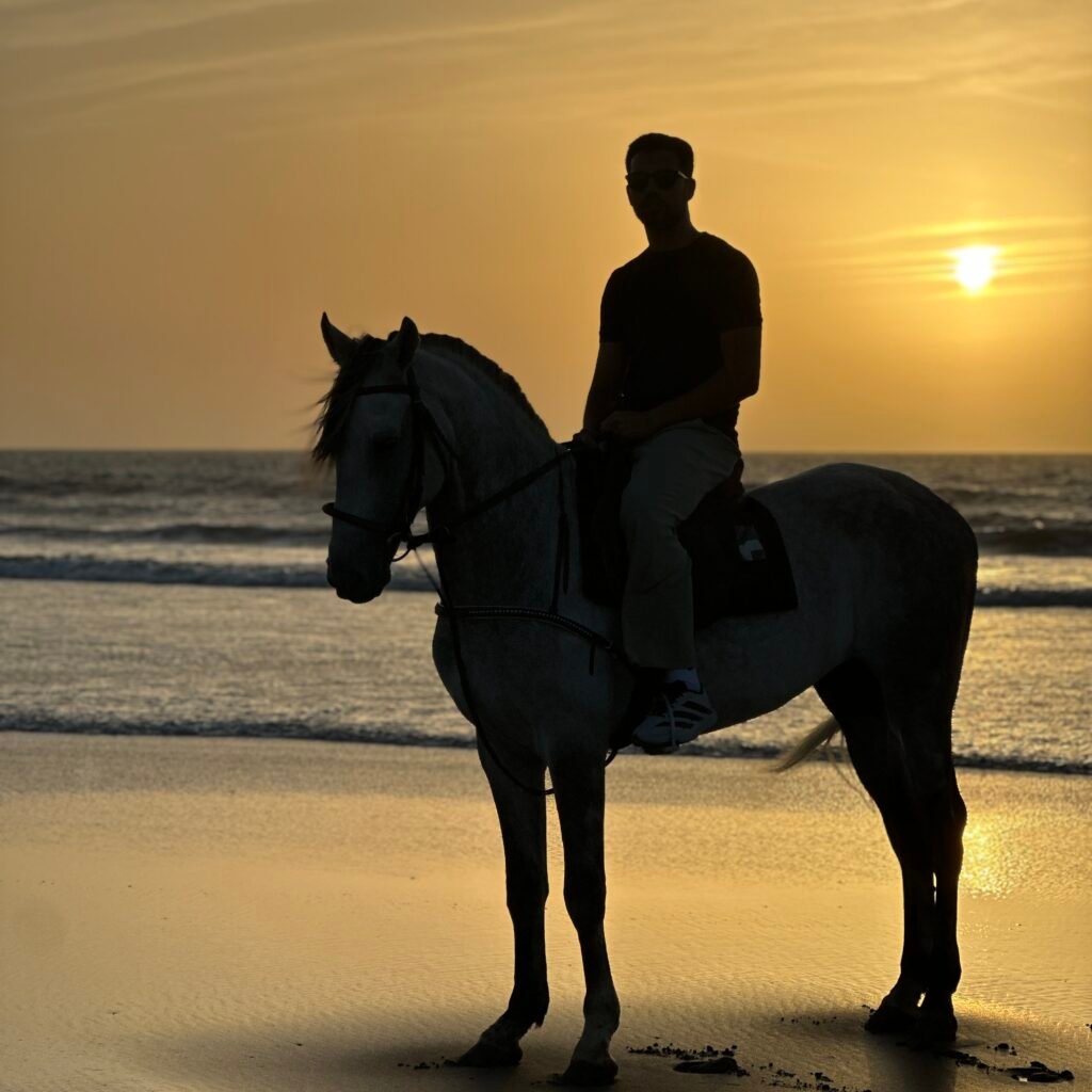 Male traveler wearing sunglasses riding a white horse along the shore during golden hour