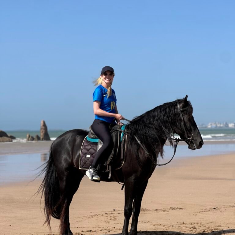 Smiling female traveler in a cap riding a black horse on a sunny beach