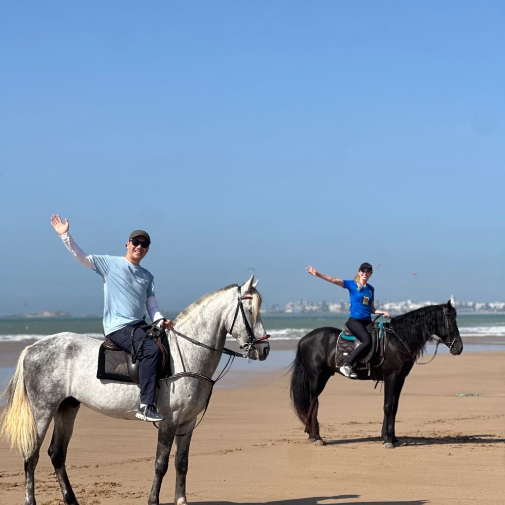 Two travelers waving while horseback riding along the beach