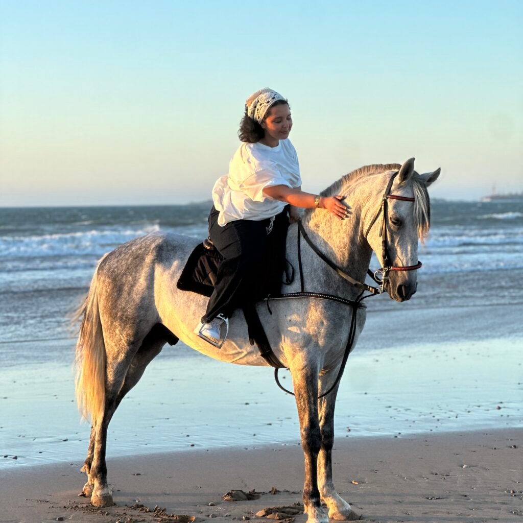 Female traveler in a white shirt and headscarf gently petting her white horse on the beach